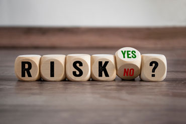 Wooden cubed dice with black letters spell out the word “risk,” followed by a cube titled on its side to show “yes” in green and “no” in red, ending with a question mark cube.