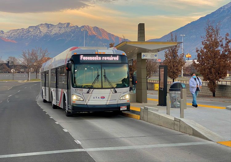 Front view of a large, silver bus with its headlights on at a BRT station in Orem, Utah. A snow-capped mountain is in the background.